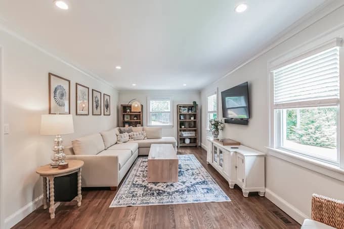 This is an image from Chameleon Painting Inc., located in New York. The photo shows a bright living room with white walls, a sectional sofa, hardwood floors, framed wall art, and natural light from multiple windows.