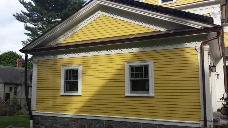 This is an image from Decolores Painting, featuring the side of a house painted in bright yellow with white trim. The stone foundation contrasts with the vivid siding and detailed trim work.