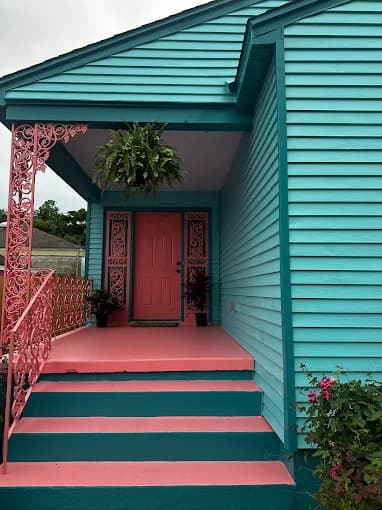 This is an image from Pelican Paint Group, located in Louisiana. The photo shows a small home with bright turquoise siding and a pink front porch, door, and trim, highlighting a bold and colorful exterior paint design.