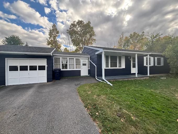 This is an image from Bates Brothers Painting LLC, located in New York. A single-story ranch home with a freshly painted dark navy blue exterior, white trim, an attached garage, and a covered side porch.