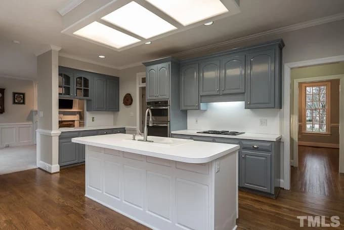 This is an image from 2 Cabinet Girls, located in North Carolina. The photo shows a spacious kitchen featuring gray refinished cabinets, a white island with a built-in sink, and warm hardwood floors creating a balanced, contemporary look.