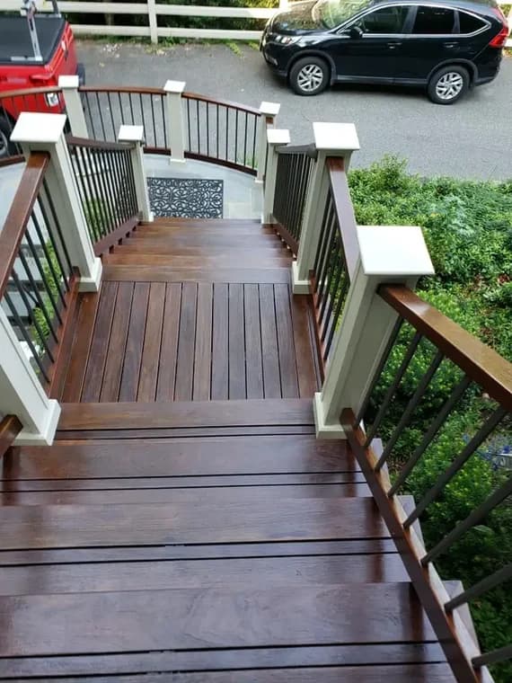 This is an image from Sam Professional Painting LLC, located in Maryland. The image shows a stained wooden staircase with railings leading down to a driveway, highlighting detailed exterior wood finishing.