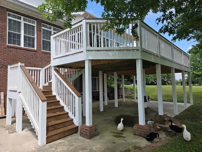 This is an image from Quick Painting LLC, located in Kentucky. The image shows a freshly painted elevated deck with white railings, wood stair treads, and support posts beneath a brick home exterior.