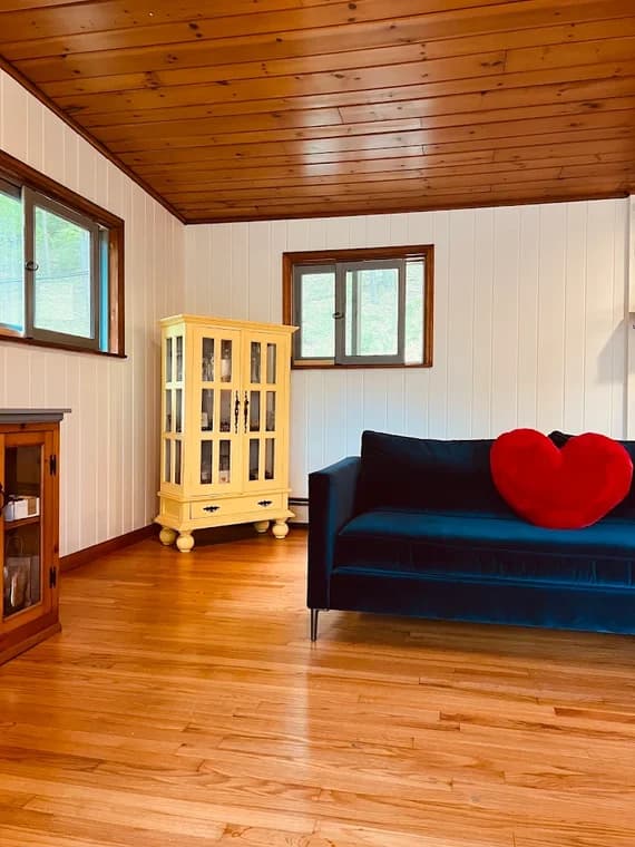 This is an image from Certified Painting, located in New York. The photo shows a bright interior room with white paneled walls, a wood-planked ceiling, hardwood floors, a blue sofa with a red heart pillow, and a yellow display cabinet.