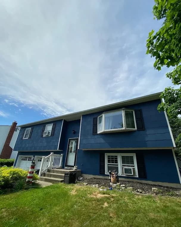 This is an image from KB Quality Painting, located in New York. The exterior photo shows a split-level home with dark blue siding, white trim, and black shutters after a fresh repaint.