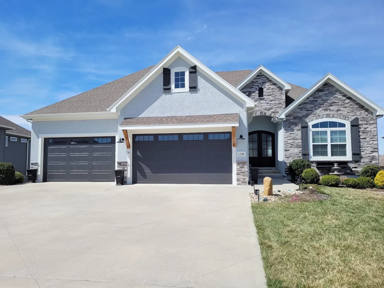 This is an image from PEMCO Painting Interior & Exterior Painter Overland Park. The image is an exterior home in Overland Park KS with fresh light blue-gray stucco, stone accents, and newly painted dark gray garage doors and shutters.