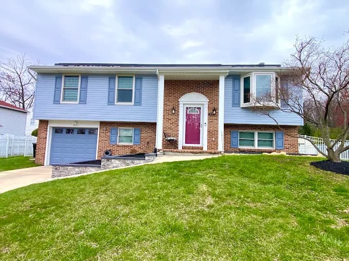 This is an image from Perfect Painters & More, located in Maryland. The photo shows a split-level home with a fresh exterior paint job featuring light blue siding, white trim, and a bold red front door accented by a brick facade.