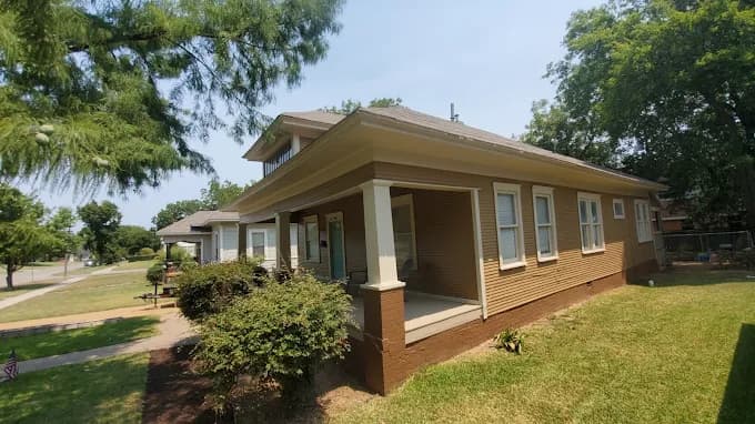 This is an image from Derheim Painting, located in Oklahoma. The photo shows a single-story home with freshly painted brown siding, cream trim, and a covered front porch surrounded by greenery.