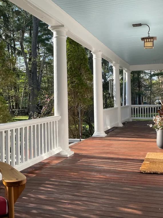This is an image from Ortolani & Sons Painting Company, located in Massachusetts. The image shows a freshly painted front porch with white columns, white railings, a light blue ceiling, and a stained wooden deck floor.