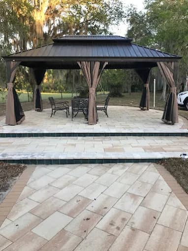 This is an image from Golden Hands Handymans company, located in Windermere, FL. The image is a custom-built outdoor gazebo with patio pavers and metal roof, featuring patio dining furniture in a landscaped backyard