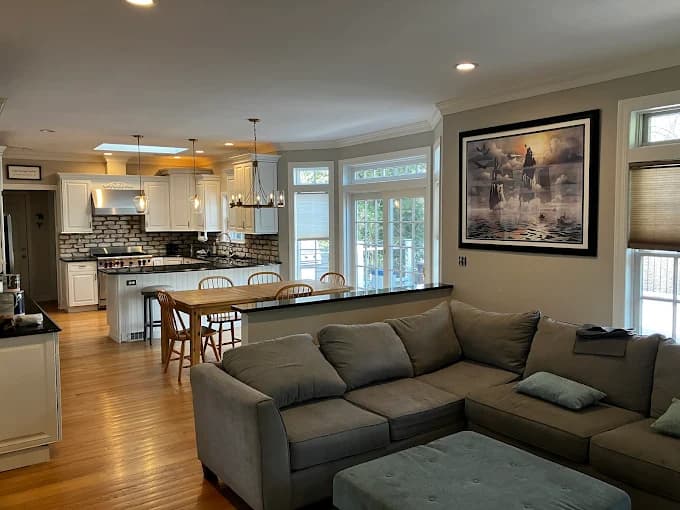 This is an image from Anthony's Painting & Decorating, Inc., located in New York. The photo shows a spacious open-concept living room and kitchen with freshly painted light-gray walls, white cabinetry, hardwood floors, and a sectional sofa.
