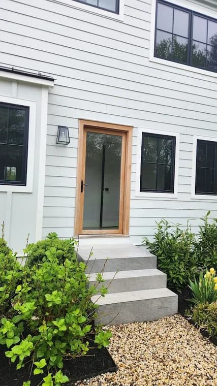 This is an image from CRD Painting Inc, located in New York. The image displays a clean white modern home exterior with black window frames and a natural wood-framed glass front door surrounded by landscaping.