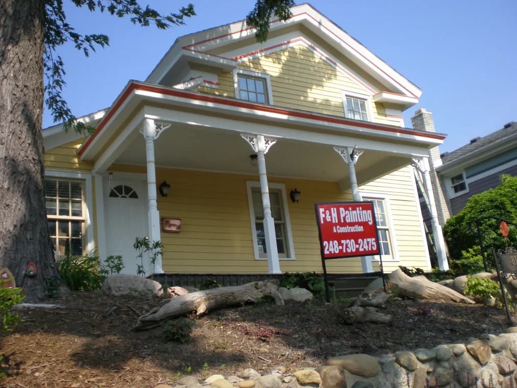 This is an image from The Exterior Painting Experts - F&H Painting company, located in Oxford, MI The image is a freshly painted yellow house with white trim and decorative porch columns, featuring an F&H Painting & Construction yard sign with phone number 248-730-2475.