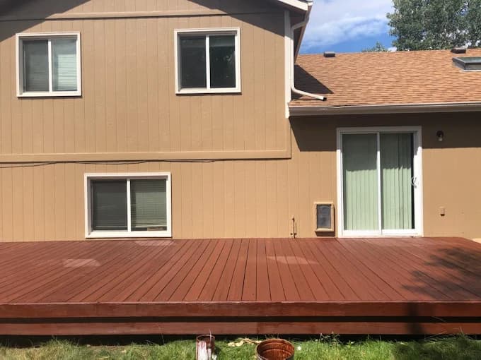 This is an image from Noble Painting company, located in Fort Collins, CO The image is a back of a beige split-level home with freshly stained redwood deck, white-trimmed windows, and sliding glass door, deck refinishing project by Noble Painting