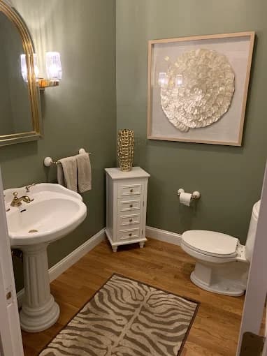 This is an image from Aurora Exterior Painting Inc., located in Massachusetts. The photo shows a small bathroom with olive green painted walls, a pedestal sink, white fixtures, wood flooring, and decorative wall art.