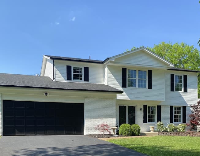 This is an image from SNL Painting Inc, located in Missouri. The image shows a freshly painted two-story home exterior with white siding, black shutters, a white brick lower façade, and a black garage door, showcasing expert exterior painting and color contrast work.