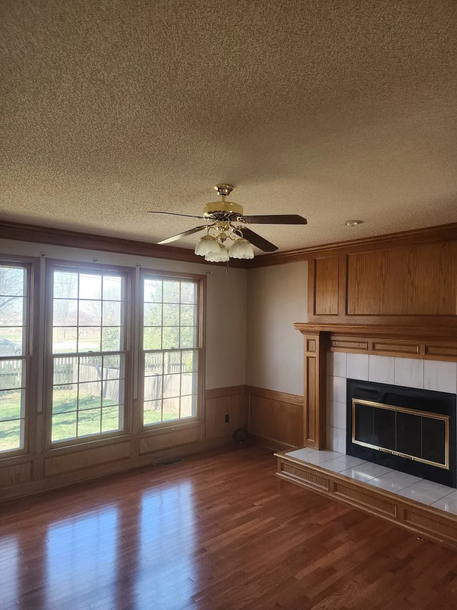 This is an image from 360 Painting of Overland Park. The image shows a living room with wood-paneled walls, large front windows, wood flooring, and a traditional fireplace framed by decorative wood trim.