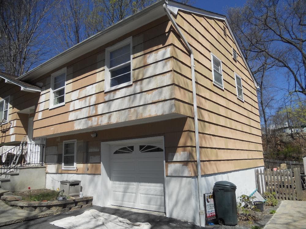 A house undergoing painting preparation, with brown siding partially primed in white, ladders leaning against the side, and a garage door below.