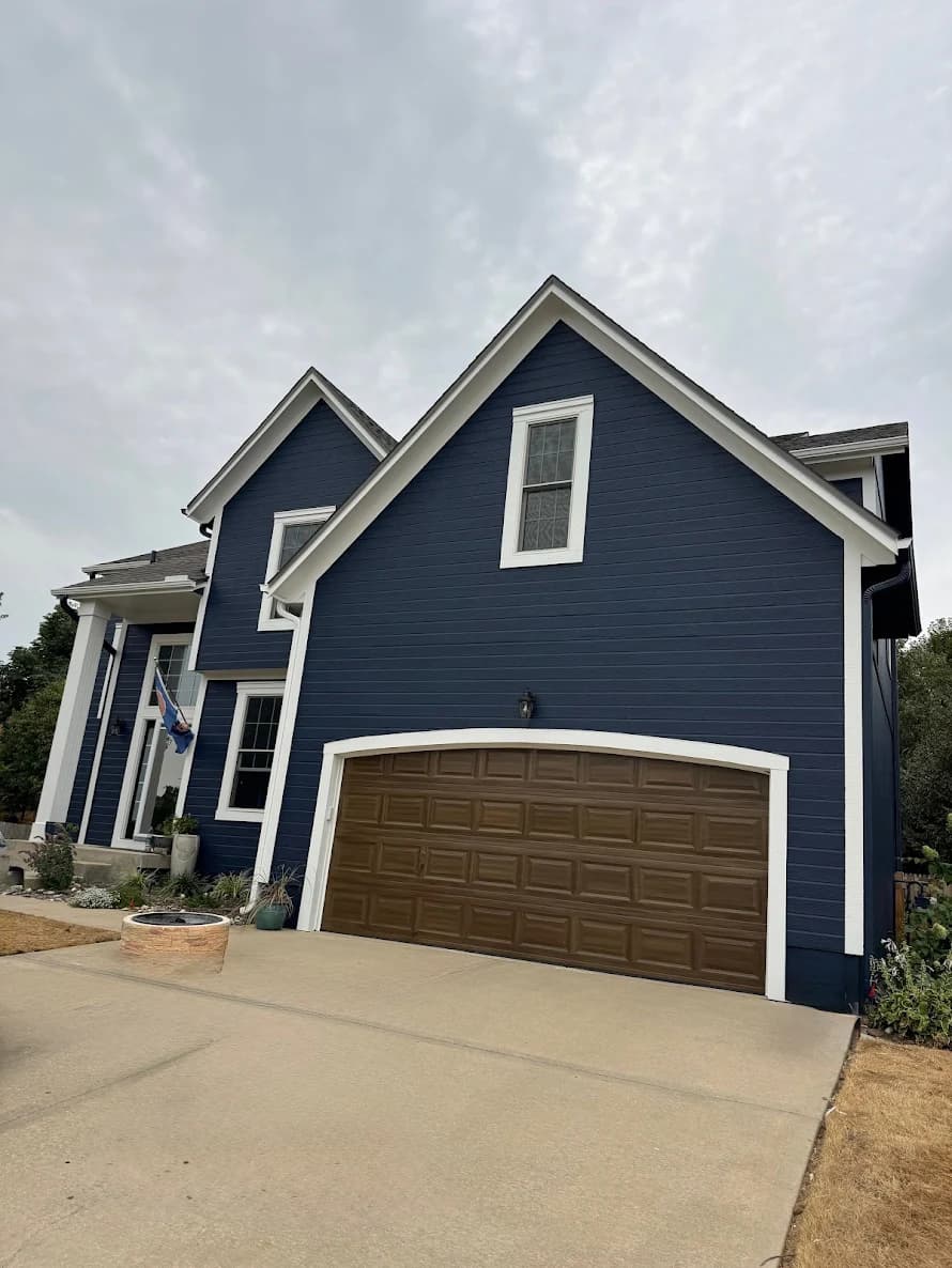 This is an image from CertaPro Painters of Shawnee Mission, KS. The image shows a freshly painted two-story home featuring deep navy blue siding, crisp white trim, and a wood-tone garage door, giving the exterior a clean and updated look.