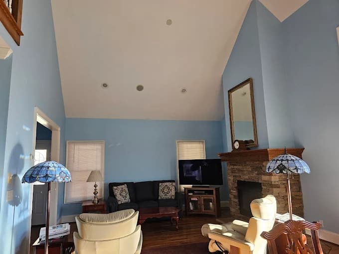 Interior painting project by Huskey's Paint & Design in Sylva, NC. The image shows a light blue living room with a vaulted ceiling, stone fireplace, and wall art.