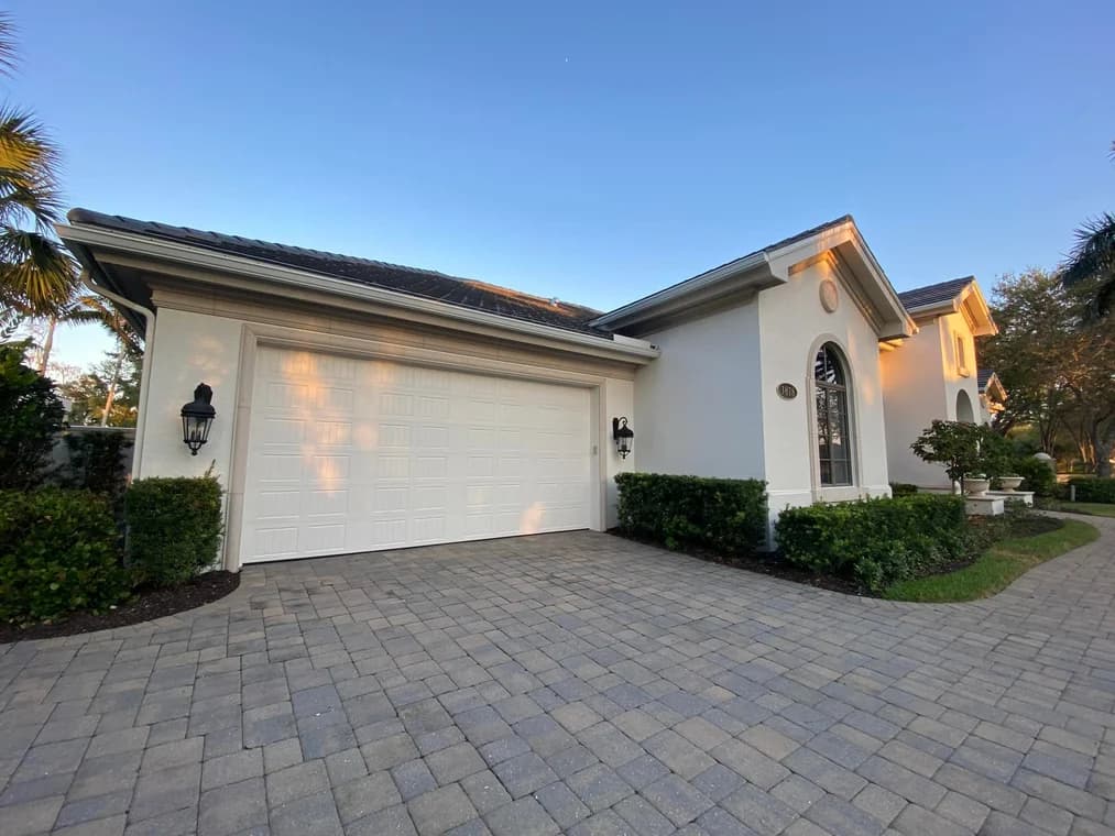 This is an image from Creative Colors And Design company, located in Naples, FL. The image is an exterior of a freshly painted white stucco home with modern garage doors, paver driveway, and manicured landscaping by Creative Colors and Design
