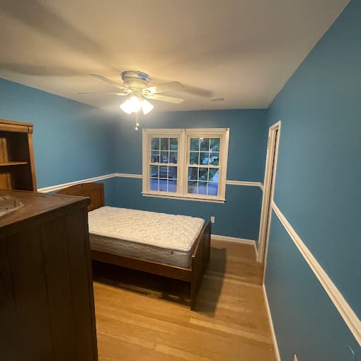 This is an image from Premier Painting Pros, located in New York. The photo shows a bedroom with blue painted walls, white trim and chair rail, hardwood flooring, and a ceiling fan above a wooden bed frame.