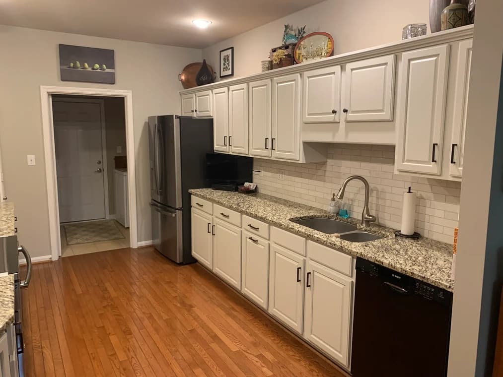 This is an image from Serious Business Painting LLC, located in Kentucky. The image shows a freshly painted kitchen interior with white cabinets, granite countertops, subway tile backsplash, stainless steel appliances, and hardwood floors.