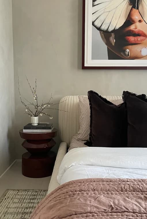 This is an image from Interior Finishing NYC, located in New York. The bedroom vignette includes a textured painted wall, framed artwork above the bed, layered bedding, and a modern side table with books and a vase.