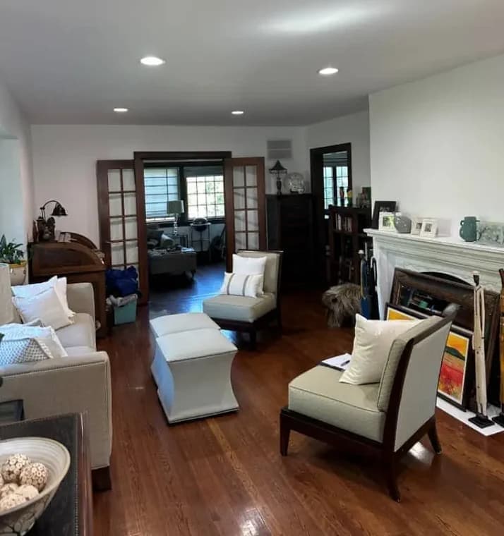 This is an image from Pier Painting Services, located in NY. The living room includes clean white walls, recessed lighting, and wood flooring with furniture arranged around a fireplace.