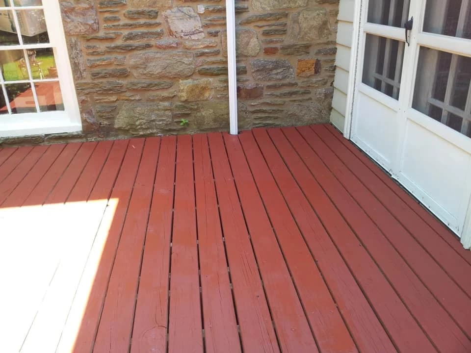 This is an image from Disbrow Painting And Handyman Services Company company, located in Levittown, PA. The image is a recently painted red wooden deck next to a stone wall.