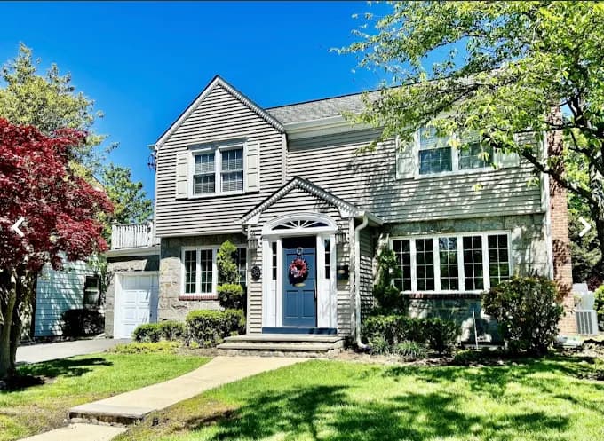 This is an image from Elite Painting Services Plus, located in New York. The exterior home image shows a two-story house with light gray siding, white trim, and a blue front door surrounded by a landscaped yard.
