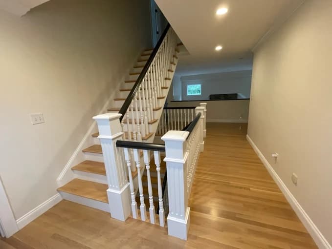 This is an image from JK Painting Service, located in Massachusetts. The photo shows a staircase with freshly painted white spindles and dark handrails, paired with polished hardwood floors for a clean, modern interior look.
