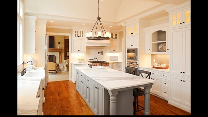 This is an image from 2 Cabinet Girls, located in North Carolina. The photo shows a bright kitchen with white cabinetry, a large gray island with marble countertops, warm wood flooring, and elegant pendant lighting over the workspace.