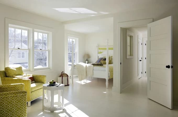 This is an image from Platinum Painting Service, located in New York. The photo shows a bright bedroom with freshly painted soft white walls, natural light from multiple windows, and coordinated furniture in light tones.