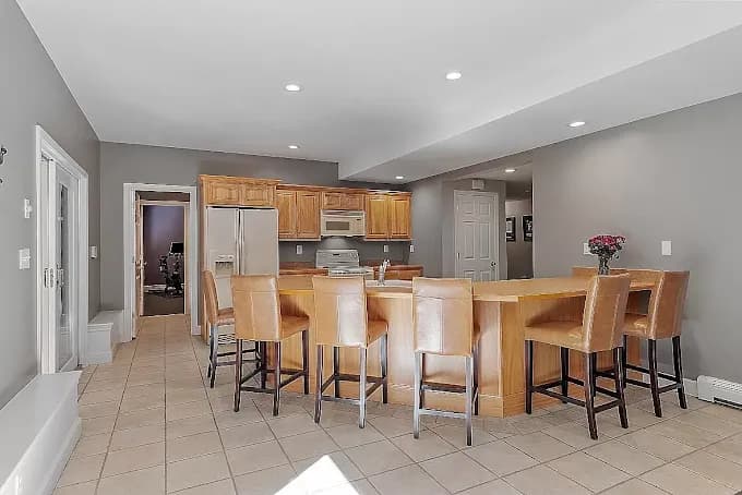 This is an image from Fresh Coat Painters of Andover, located in Massachusetts. The photo shows a spacious kitchen with gray painted walls, wooden cabinetry, a large breakfast bar with leather chairs, and tile flooring.