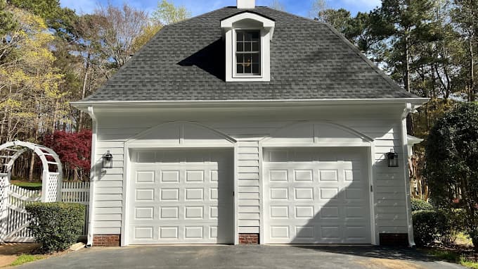 Exterior painting project by Triangle Pro Painting in Raleigh, NC. The image shows a detached white garage with two doors, black shingles, and trees.