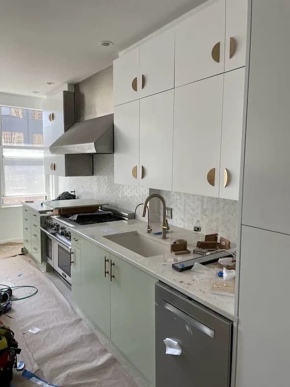This is an image from Nyca Contractors, LLC, located in New York. The image shows a kitchen under construction with white upper cabinets, light green lower cabinets, brass hardware, and a white textured tile backsplash.
