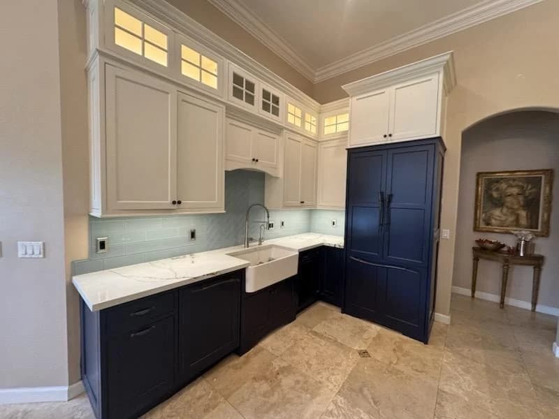 This is an image from Big Family Painting Co  company, located in Boca Raton, FL. The image is a two-tone kitchen cabinets with white uppers and navy blue lowers, featuring a farmhouse sink, marble countertops, and under-cabinet lighting in a luxury home kitchen