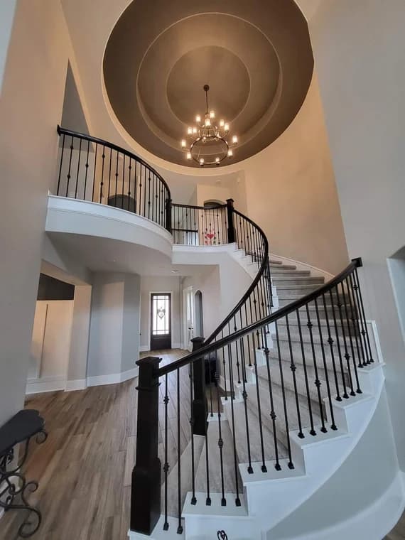 "This is an image from Reyes Painting Co., located in Texas. The photo captures a grand curved staircase with black spindles, white trim, and a multi-tiered dome ceiling featuring a hanging chandelier."