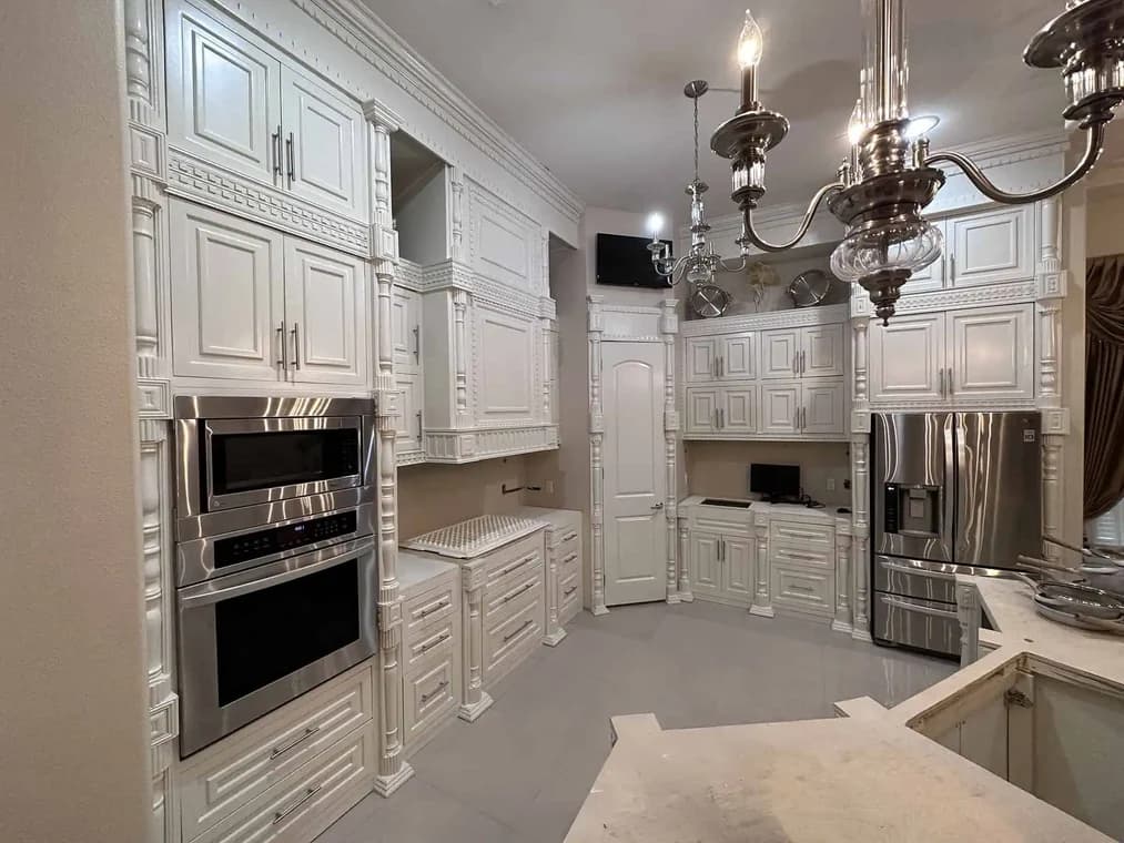 "This is an image from Armored Painting, located in Texas. The photo shows a detailed white kitchen renovation with ornate cabinetry, stainless steel appliances, and polished chandeliers above the workspace."