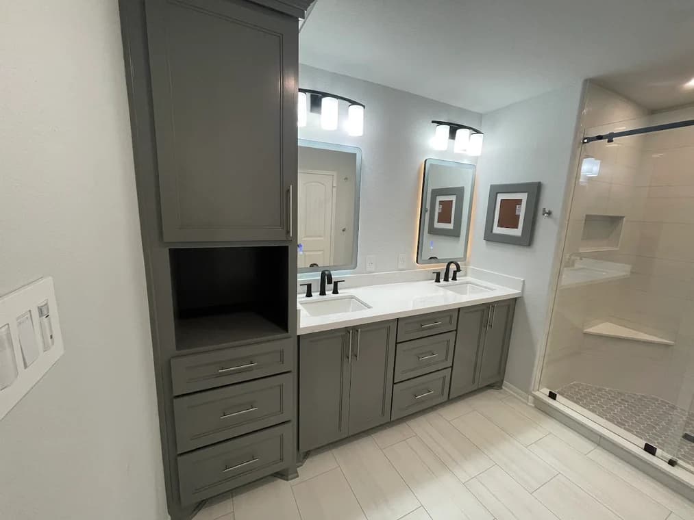 "This is an image from Lee House Paint & Repair, located in Texas. The photo shows a remodeled bathroom with gray cabinetry, dual sinks, modern lighting, and a glass-enclosed shower."