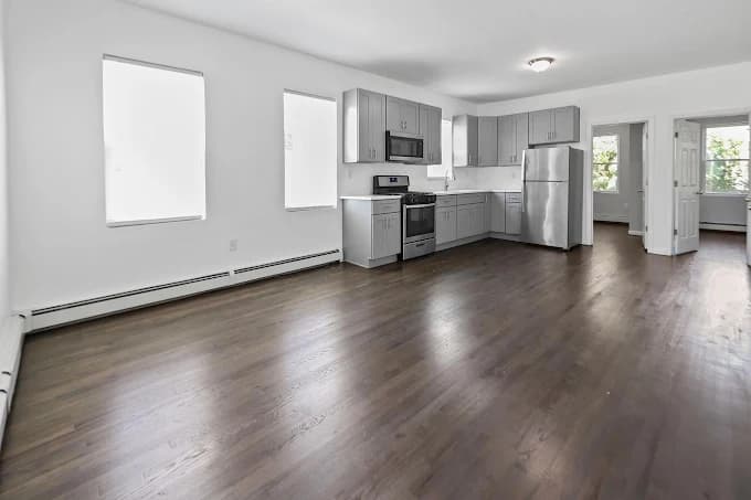 This is an image from Premier Painting Pros, located in New York. The photo shows a bright, freshly painted open-concept kitchen and living area with white walls, gray cabinetry, stainless steel appliances, and dark hardwood flooring.