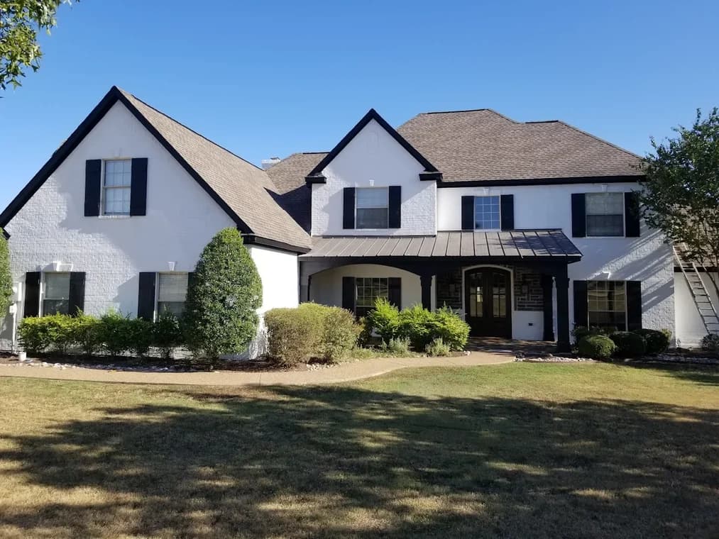 "This is an image from Texas Star Painting, located in Texas. The exterior image shows a two-story white brick home with black shutters, a dark metal porch roof, and neatly trimmed landscaping."