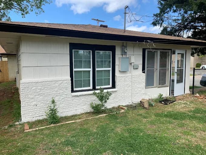 This is an image from Cardinal Painting, located in Oklahoma. The photo shows a freshly painted small home exterior with white siding, a brick lower section, and black trim around the windows and doors.