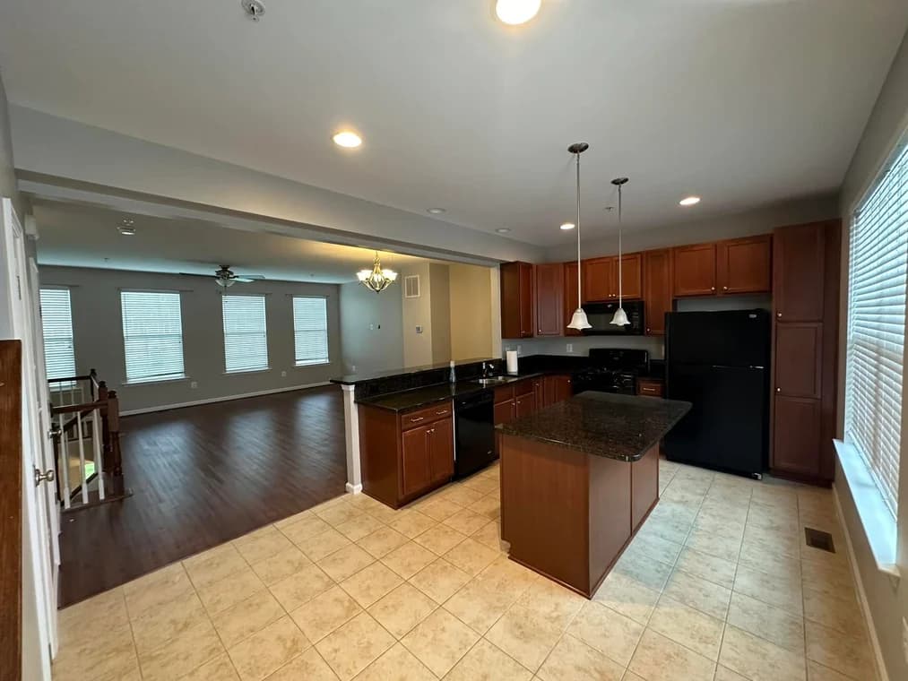This is an image from DJ’s Painting & Powerwashing, located in Maryland. The image shows a kitchen and living area with dark wood cabinets, granite countertops, tile flooring, and an open floor plan leading into a bright living room.