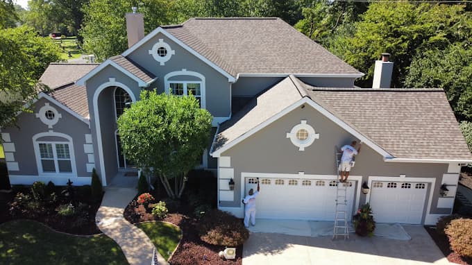 This is an image from Kennedy Painting Interior & Exterior, located in Indiana. The image shows a large residential home exterior being professionally painted with neutral gray siding, white trim, and multiple painters working on ladders to complete an exterior repaint.