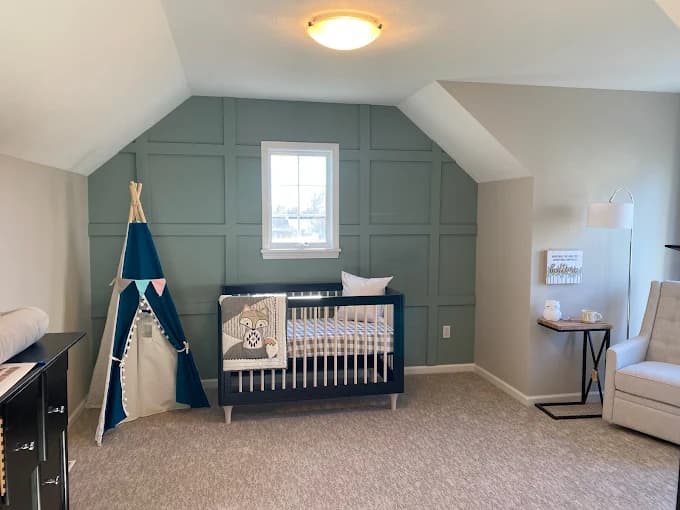 This is an image from Monarch Painting, located in New York. The photo shows a nursery with a green accent wall featuring decorative paneling, a crib, a small play tent, and neutral-toned furnishings.