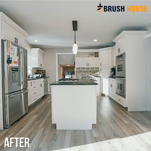 This is an image from Brush House Painting & Flooring Services of North Jersey, located in New Jersey. The image shows a renovated white kitchen with a large center island, stainless steel appliances, recessed lighting, and wood plank flooring.