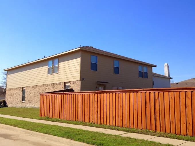 This is an image from Big Al's Painting, located in Texas. The exterior photo shows a two-story beige and brick home with a freshly painted wooden fence under a clear blue sky.
