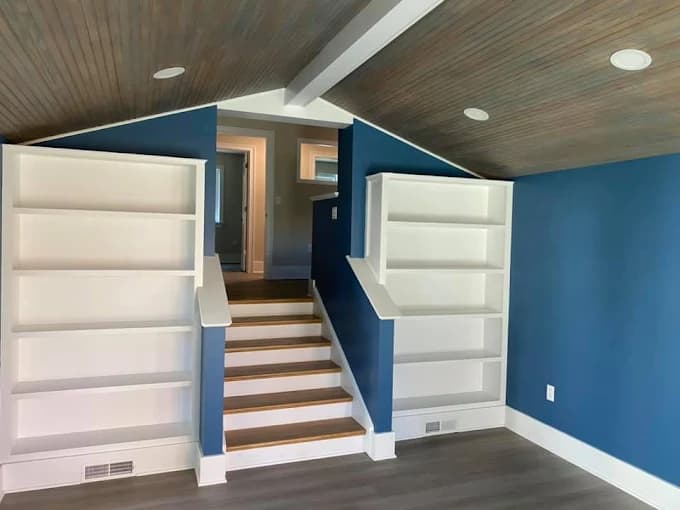 This is an image from JT’s Painting 518 LLC, located in New York. The image shows a finished interior space with deep blue painted walls, custom built-in shelving, white trim, and a wood-accent ceiling.
